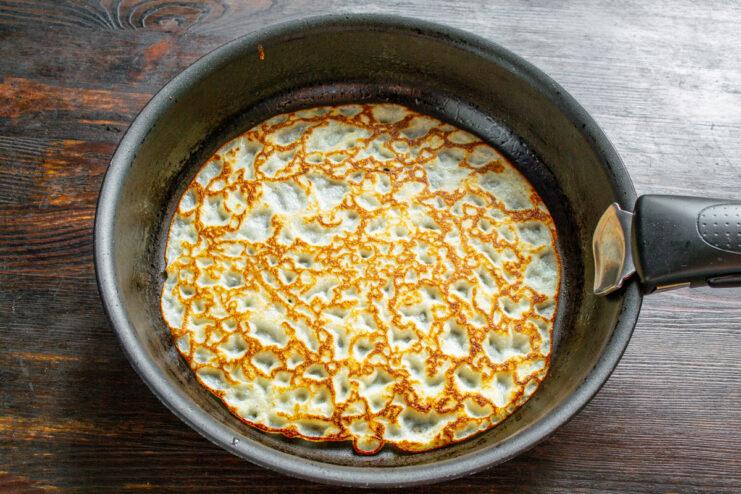 Flipping the thin pancake to cook it to golden perfection. A thin, golden-brown pancake being flipped in a black skillet with a spatula.