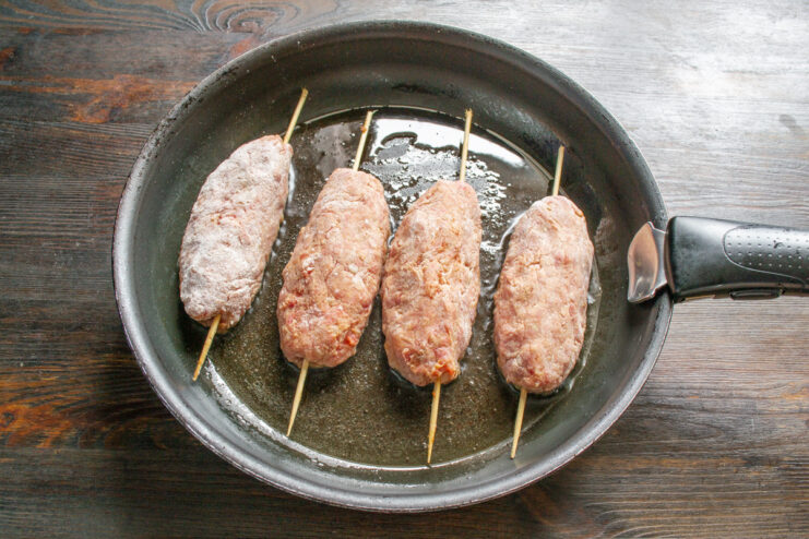 Placing Skewered Sausages onto a Hot Skillet Placing four beef sausages on skewers into a hot, oiled skillet.
