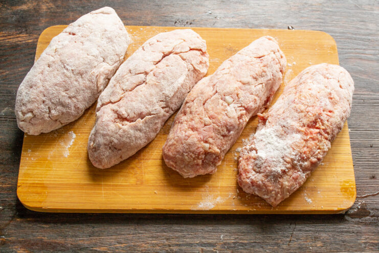 Shaping and Dredging the Beef Sausages Four raw beef sausages shaped and coated in flour on a cutting board.