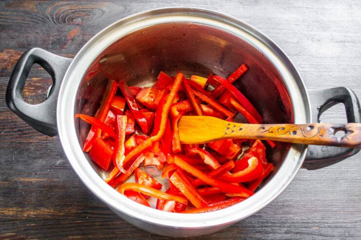 Adding sliced bell peppers to the pot with tomatoes. A pot containing diced tomatoes with freshly sliced red bell peppers being added.