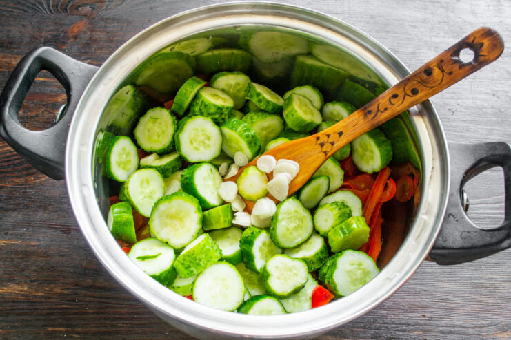 Adding fresh cucumbers and garlic to the simmering vegetables. Sliced cucumbers and garlic being added to a pot of simmering tomatoes and peppers.