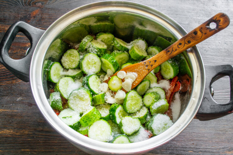 Seasoning the Georgian cucumber salad with salt and sugar. Adding a spoonful of sugar and salt to the vegetable mixture in the pot.