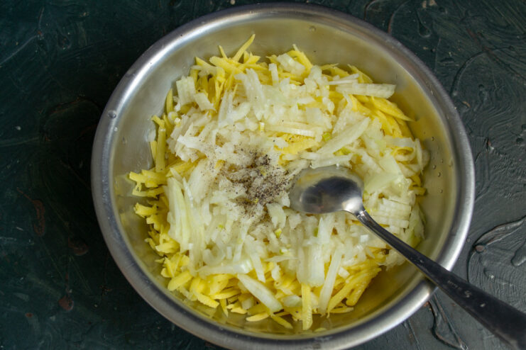 Seasoning the potato and onion mixture well is key to a flavorful draniki. A bowl of grated potatoes and chopped onions being seasoned with salt and pepper.