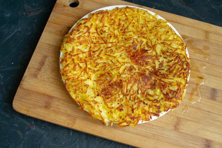 A clever trick to flip the large draniki without breaking it, similar to a Spanish tortilla. Flipping a large potato pancake from a skillet onto a plate over a cutting board.
