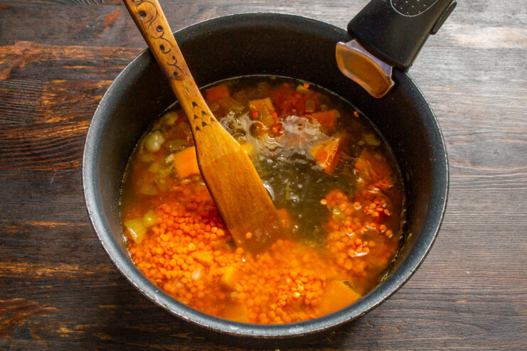 Pouring chicken broth into the pan to simmer the vegetables and lentils. Chicken broth being poured into a pan with pumpkin, lentils, and onion.