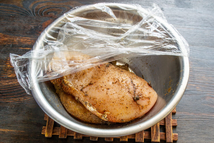 A hand rubbing spices into a raw chicken breast fillet in a glass bowl.