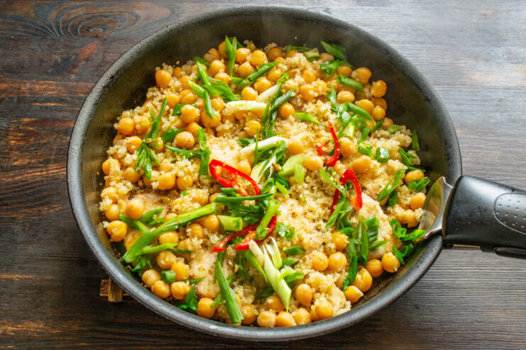 Chopped scallions and chili rings being sprinkled over the chicken, quinoa, and chickpeas in the skillet.