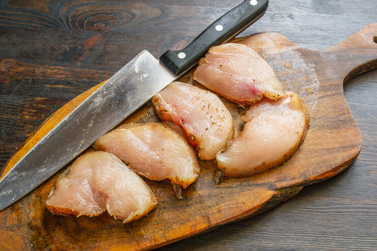 A hand with a knife slicing a marinated chicken breast on a cutting board.