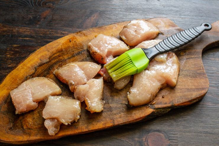 A silicone brush applying oil to sliced raw chicken breast pieces.
