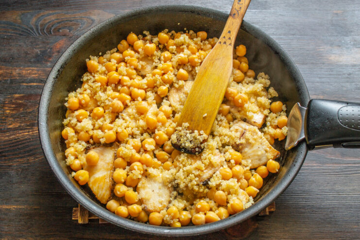 Cooked quinoa being added to the skillet with chicken and chickpeas.