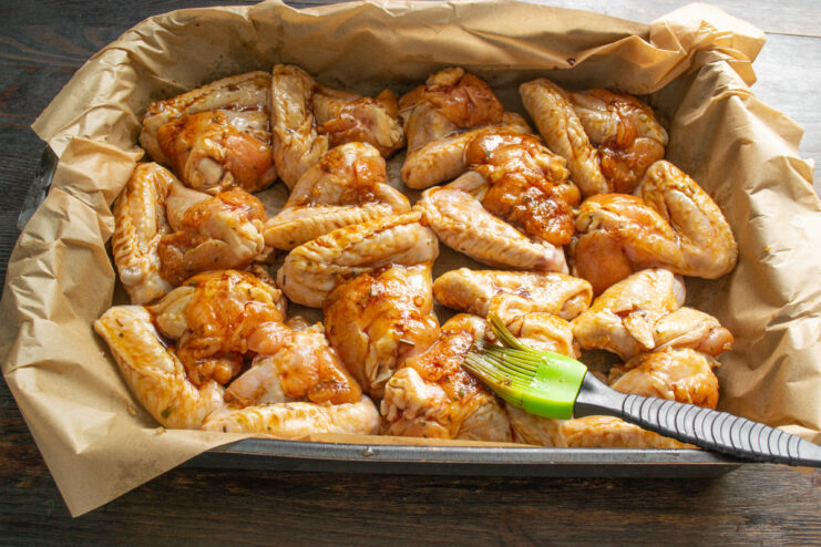 How to Get a Perfectly Sticky Glaze on Your Wings A hand using a silicone pastry brush to coat chicken wings with marinade on a baking sheet.