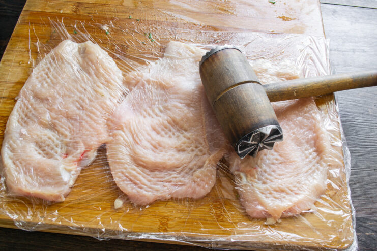 Tenderizing the Chicken Schnitzels with a Meat Mallet Pounding chicken cutlets covered with plastic wrap using a meat mallet to tenderize them.