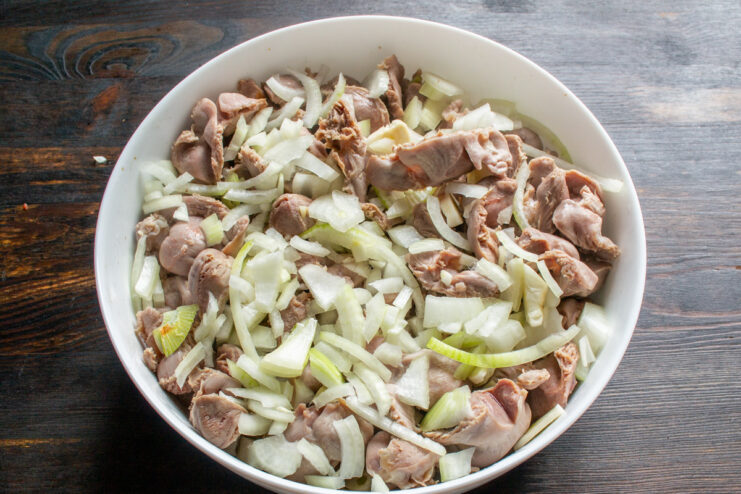 Adding finely chopped onions to the gizzards. Finely chopped raw onion being added to the chicken gizzards in a baking dish.