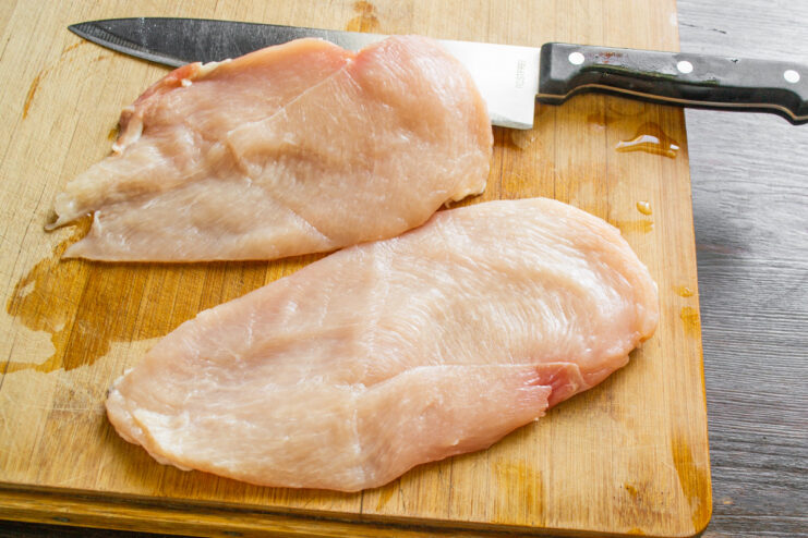 How to Butterfly a Chicken Breast for a Roulade A hand holding a sharp knife slicing a large raw chicken breast in half horizontally on a cutting board.