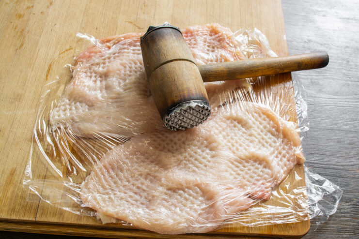 Pounding Chicken Thin for an Evenly Cooked Roulade A chicken breast covered with plastic wrap being pounded thin with a metal meat mallet.