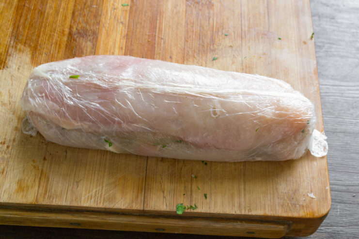 How to Tightly Roll the Chicken Roulade Using Plastic Wrap A chicken breast with filling being carefully rolled into a tight log using plastic wrap to help shape it.
