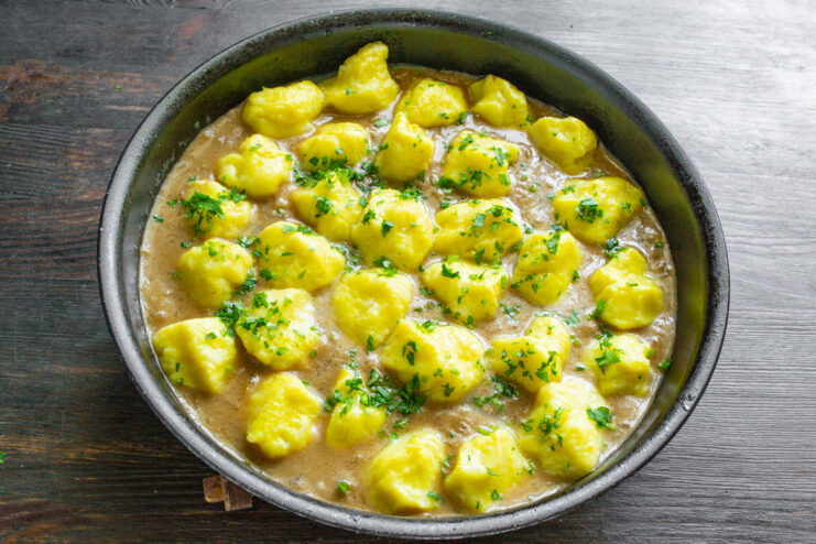 Finishing the dish by combining dumplings with sauce and herbs. Cooked lazy potato varenyky being gently mixed into the creamy mushroom sauce in a pan, garnished with herbs.