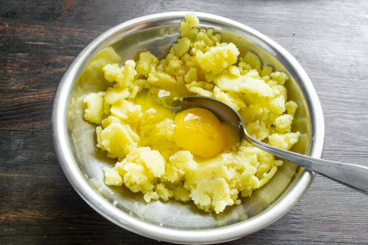 Combining mashed potatoes with egg and salt to start the dough. A glass bowl with mashed potatoes, a cracked egg on top, and a pinch of salt being added.