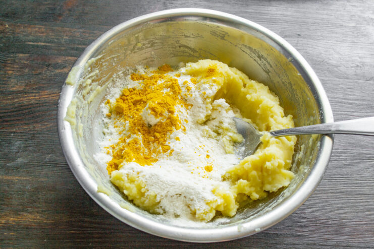 Adding flour and turmeric for color and flavor to the potato dough. Mixing mashed potatoes, egg, butter, flour, and turmeric in a bowl to form the dough for lazy varenyky.