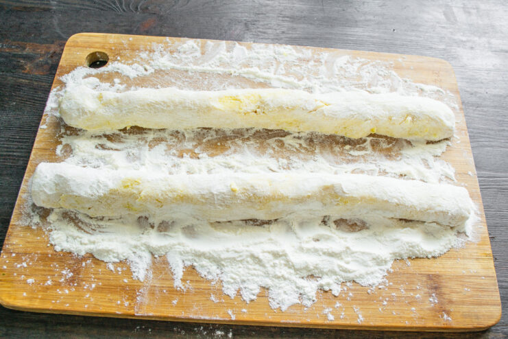 Shaping the potato dough into logs before cutting. Two long logs of potato dough on a floured wooden board, ready to be cut into dumplings.