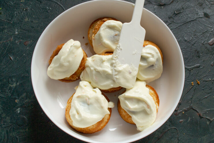 Glazing the Warm Lemon Buns A hand using a spatula to spread a thick, creamy white glaze over warm lemon buns.