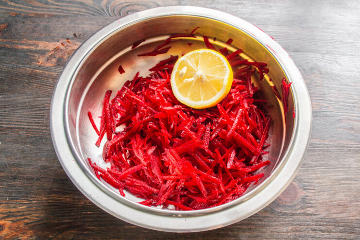 Adding lemon juice not only adds flavor but also helps preserve the vibrant color of the beets. Grated beets in a glass bowl being mixed with freshly squeezed lemon juice.