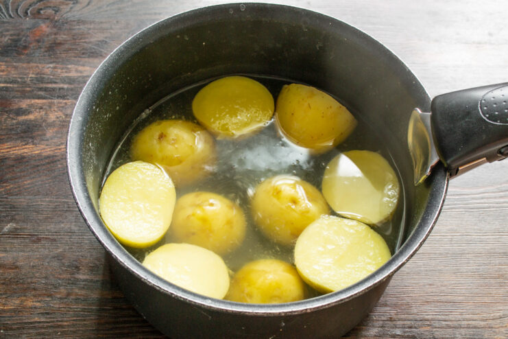 Halving new potatoes and placing them in boiling water to par-cook. Halved new potatoes in a pot, being covered with water for boiling.