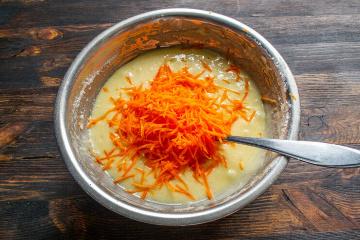 Adding Freshly Grated Carrots to the Muffin Batter Finely grated carrots being added to the smooth muffin batter in a glass bowl.