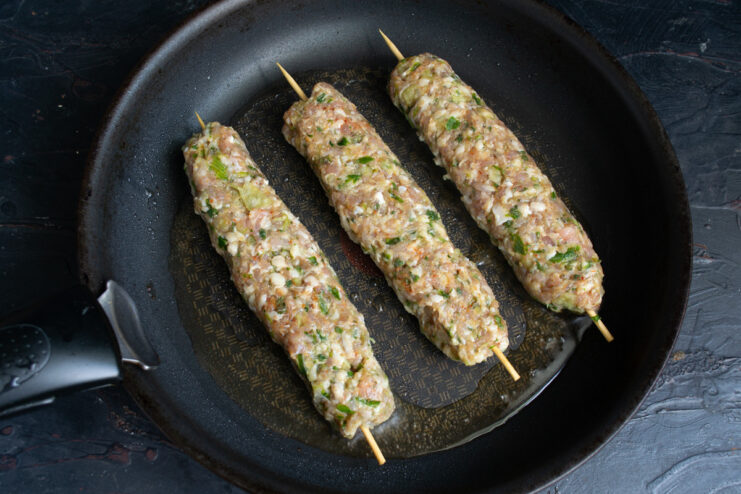 Cooking the Egyptian meat kebabs in a hot skillet. Raw meat kebabs on skewers being placed into a hot frying pan.