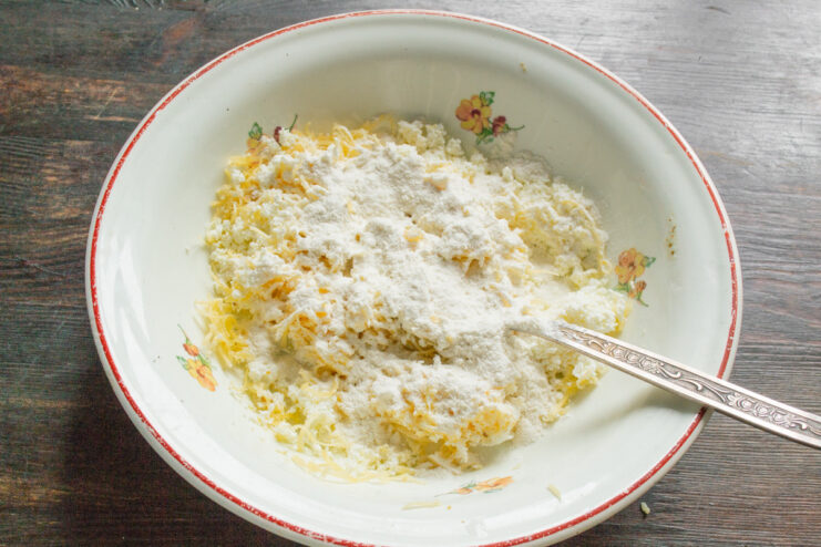 Adding Flour to the Syrniki Mixture - Just enough to bind the ingredients. A hand adding a spoonful of white flour to the bowl with syrniki ingredients.