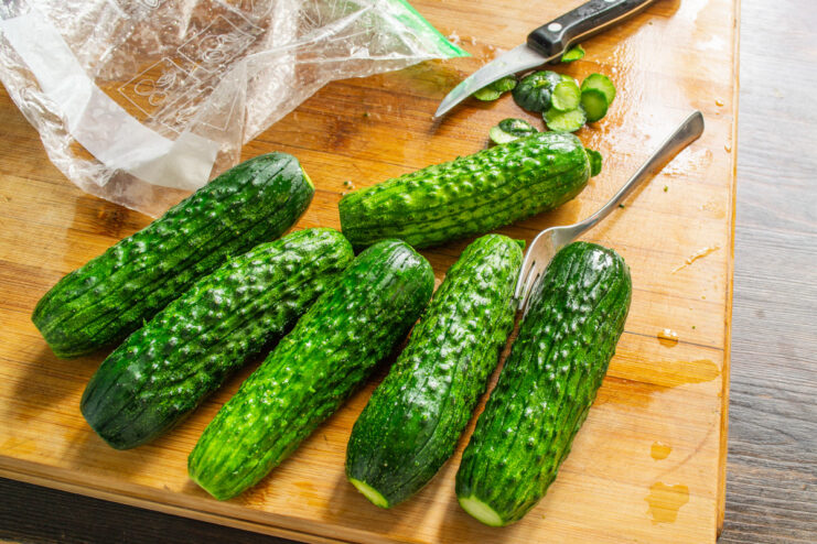 Scoring the cucumbers helps the brine penetrate faster for quick pickling. A hand holding a fork, scoring the skin of a green cucumber on a cutting board.
