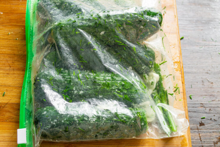 Shaking the bag ensures all ingredients are evenly distributed. Hands shaking a sealed ziplock bag containing cucumbers, dill, and salt.