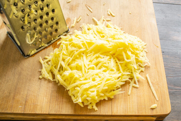 Grating Potatoes for Vegetable Fritters Peeled raw potatoes being grated on a coarse box grater into a bowl.