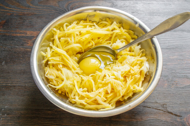 Binding the Fritter Batter with an Egg A whole egg cracked into a bowl with grated potatoes and onions.