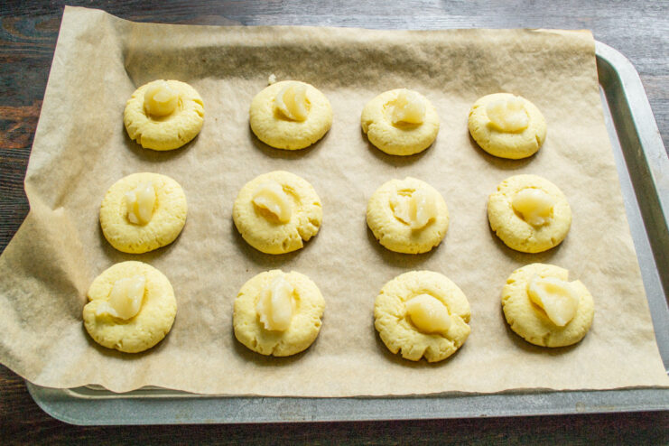 Adding a sweet honey filling to the center of each cookie. Partially baked cookies on a baking sheet with honey being added to the center indentations.