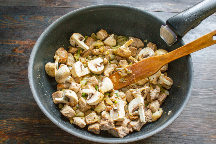 Adding sliced mushrooms to the pan with meat and onions. Sliced champignon mushrooms being added to the pan with cooked pork and onions.