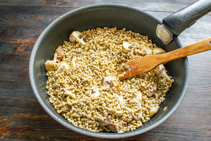 Layering the rinsed pearl barley over the meat and mushrooms. Rinsed pearl barley being spread evenly over the meat and mushroom mixture in a pan.