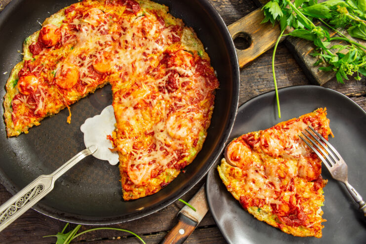 A whole skillet zucchini pizza with sausages, topped with melted cheese and herbs, served on a wooden board.