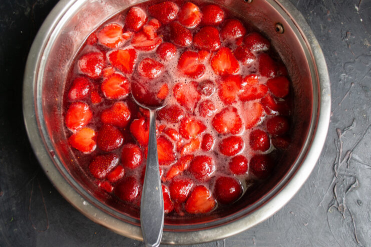 Halved strawberries being added to a saucepan with bubbling sugar syrup.
