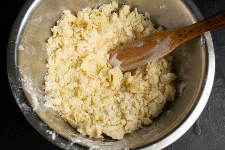 Hands rubbing butter and flour together in a bowl to create a sandy, crumb-like texture for the dough.