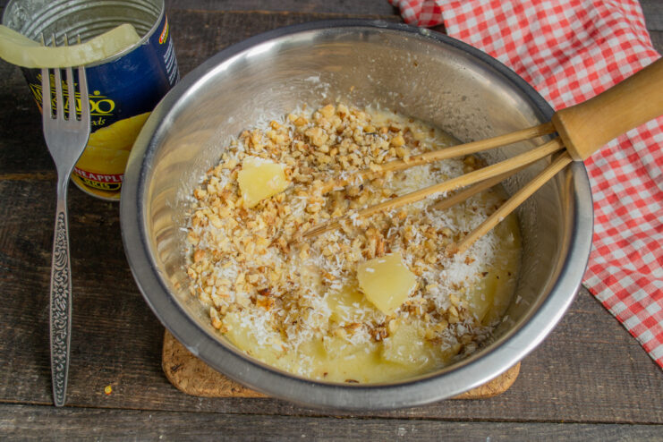 Step 4: Combining Wet Ingredients with Pineapple, Nuts, and Coconut A bowl with egg mixture, with shredded coconut, chopped walnuts, and diced pineapple being added.