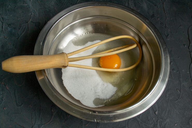 Step 1: Combining sugar, salt, and an egg in a bowl for the dough. A white bowl containing sugar, salt, and a cracked egg, ready for mixing the bun dough.