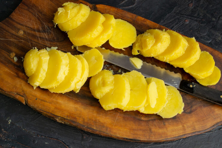 Slicing boiled potatoes for the salad base. Close-up of boiled and peeled potatoes being sliced into uniform rounds on a cutting board.