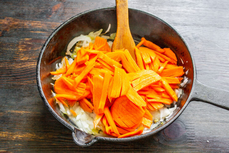 Adding shredded carrots to the skillet with onions and garlic. Shredded carrots being added to the sautéed onions and garlic in a skillet.