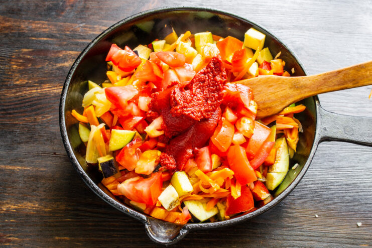 Adding rich tomato paste to the vegetables for a deeper flavor. A spoonful of tomato paste being added to the vegetables in the skillet.