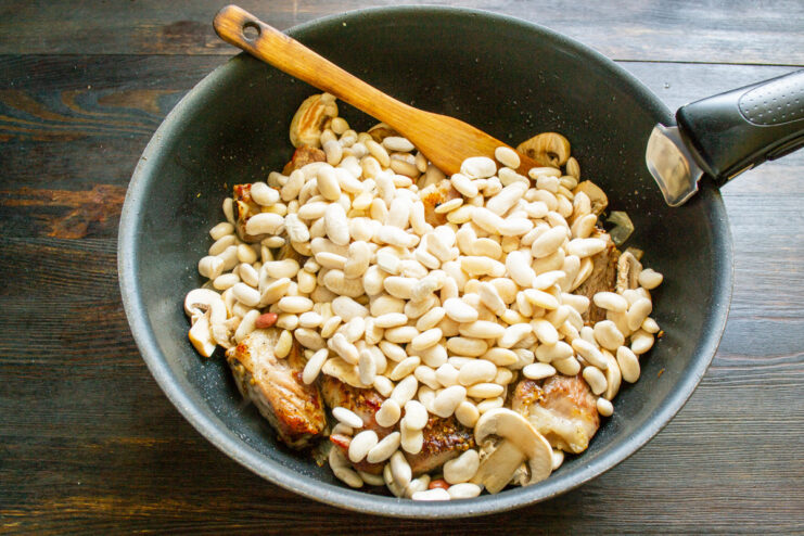 Combining the soaked beans with the seared ribs and aromatics. Soaked white beans being added to a skillet with pork ribs and vegetables.