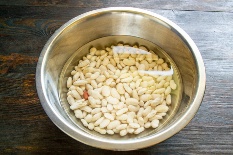 First step: Soaking dry white beans overnight to soften them for the stew. A bowl of dry white beans being soaked in clear water.