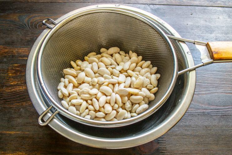 Rinsing the soaked beans is an important step to prepare them for cooking. Soaked white beans being rinsed in a metal sieve under running water.