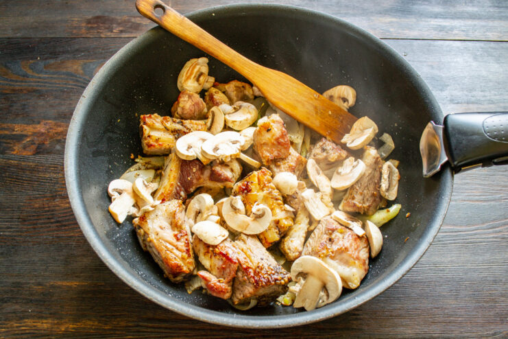 Adding sliced mushrooms to build another layer of savory flavor. Thinly sliced champignon mushrooms being added to a skillet with other ingredients.