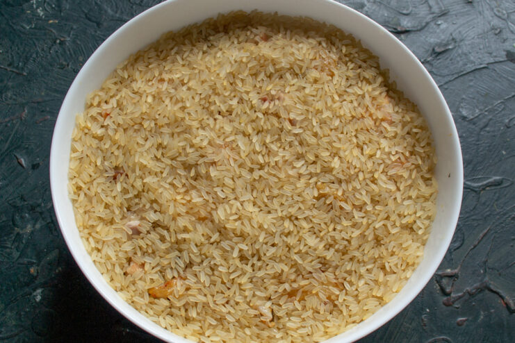 Creating an even layer of rice over the chicken and vegetables. Long-grain parboiled rice being spread evenly over the chicken and vegetables in a baking dish.